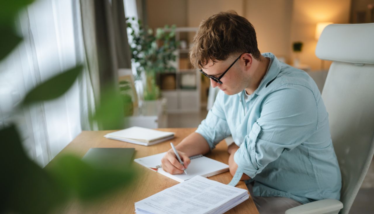 Photo of young student sitting on an office chair and writing at a desk in front of a window in a bright room