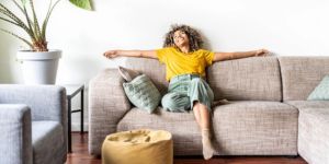 A woman in a yellow shirt and green pants relaxes with arms outstretched on a beige sofa in a bright, cozy living room. Decorative pillows and a large potted plant add comfort and style to the space.
