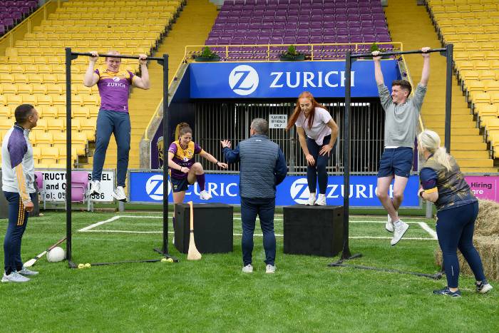A group of people are participating in a fitness challenge on a grassy field inside a stadium. Two individuals are doing pull-ups on metal bars, while others, including a coach or judge, are observing and encouraging them. There are black platforms, sports equipment like a hurley and football, and hay bales on the field. The stadium seating is yellow and purple, and Zurich branding is prominently displayed in the background.